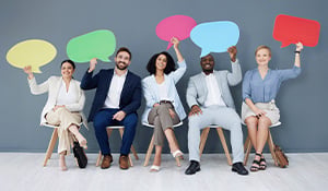 A diverse group of five professionally dressed individuals sitting on wooden chairs against a gray background, each holding a colorful speech bubble cutout above their heads.