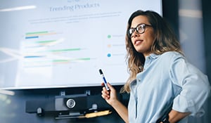 Woman standing in front of a large screen displaying a presentation with charts, holding a marker and pointing toward the screen in a meeting room.
