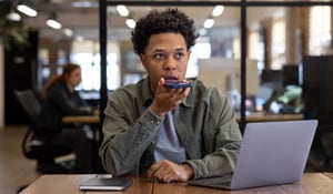 Person sitting at a desk in a modern office, speaking into their smartphone using the voice command feature.