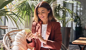 Professional woman in a rust-colored blazer smiling while using her smartphone, seated in a modern, sunlit space surrounded by large green plants, with a notebook in her lap.