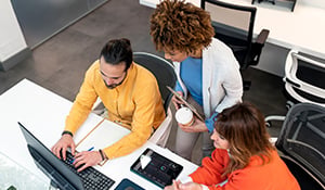 Three colleagues collaborating at a desk, reviewing information on a laptop together in a modern office setting.