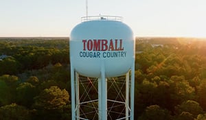 A large white water tower with red and blue lettering that reads "TOMBALL COUGAR COUNTRY," standing tall above a forested landscape at sunset. 