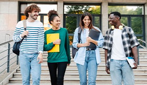 Four smiling students walk down campus steps, chatting and carrying books and backpacks.