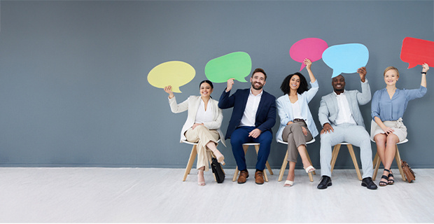 A diverse group of five professionally dressed individuals sitting on wooden chairs against a gray background, each holding a colorful speech bubble cutout above their heads.