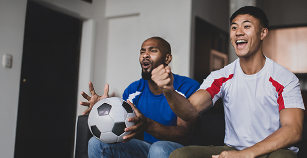 Two men sitting on a couch excitedly watching a soccer game, one holding a soccer ball while both cheer and smile.