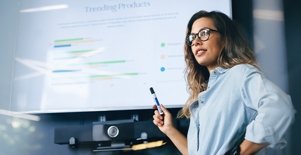 Woman standing in front of a large screen displaying a presentation with charts, holding a marker and pointing toward the screen in a meeting room.
