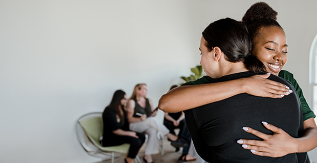 Two women warmly hugging in the foreground, while a small group of people sit and talk together in the background against a light-colored wall.