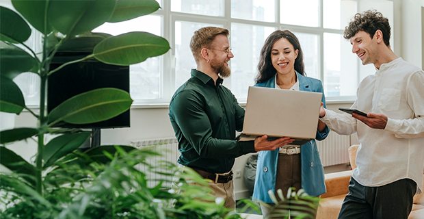 Three coworkers standing together in a bright, plant-filled office, reviewing content on a laptop while discussing ideas.