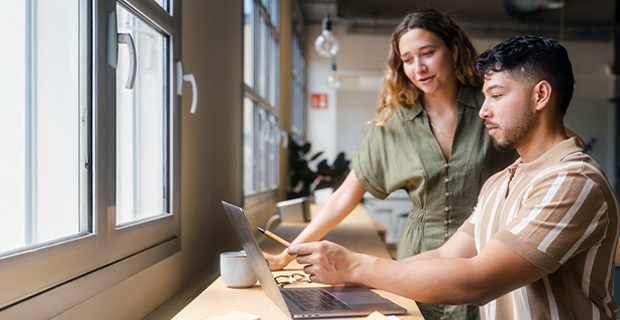 Two colleagues working together in an office; a man seated at a desk using a laptop while holding a pencil, with a woman standing beside him, both focused on the screen.