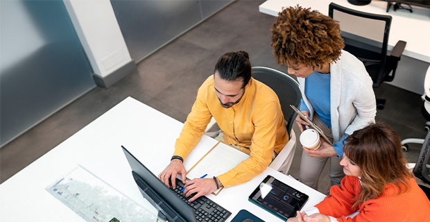 Three colleagues collaborating at a desk, reviewing information on a laptop together in a modern office setting.