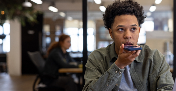 Person sitting at a desk in a modern office, speaking into their smartphone using the voice command feature.