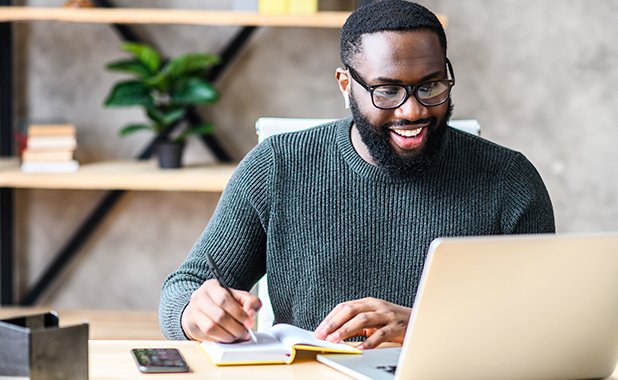 African American man looks at his laptop while actively writing in a notebook