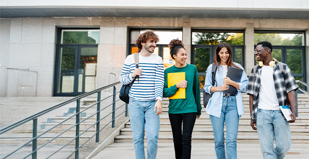 Four smiling students walk down campus steps, chatting and carrying books and backpacks.